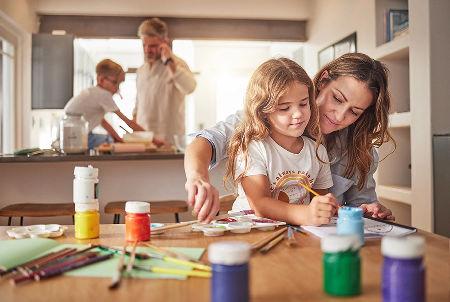 Madre e hija realizando una actividad de pintura creativa mientras el padre y el hijo colaboran al fondo en un ambiente de aprendizaje familiar
